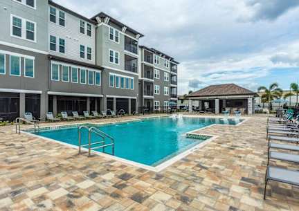 A large swimming pool surrounded by a brick patio and lounge chairs in front of a multi-story apartment building.