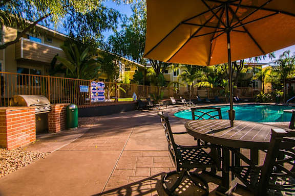A patio with a table and chairs under an umbrella with a pool in the background.