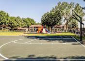 Thumbnail 4 of 26 - Apartments in Aurora, CO for Rent - Copper Flats - Basketball Court Near Grass Field and Playground with Jungle Gym, and Surrounding Trees.