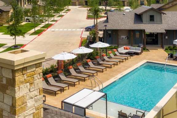A pool area with lounge chairs and umbrellas in front of a residential area.