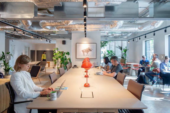 A woman in a white shirt is working on her laptop in a modern office with a long table and chairs.