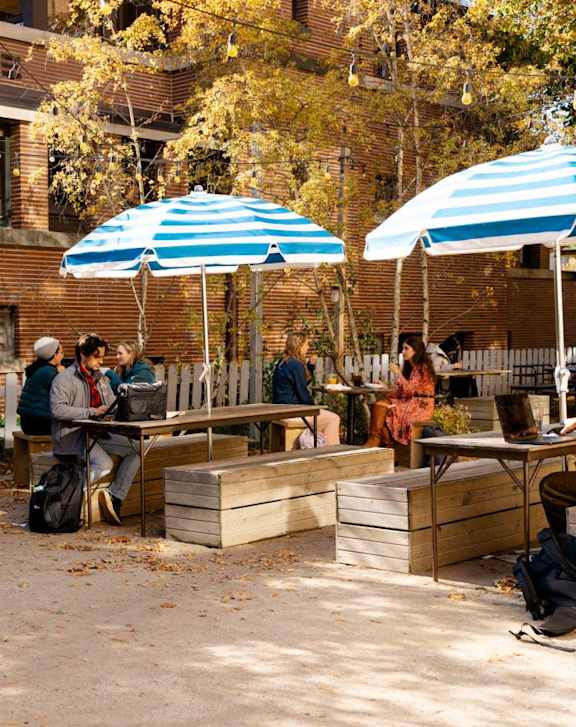 A group of people are sitting and standing on wooden benches under blue and white striped umbrellas.