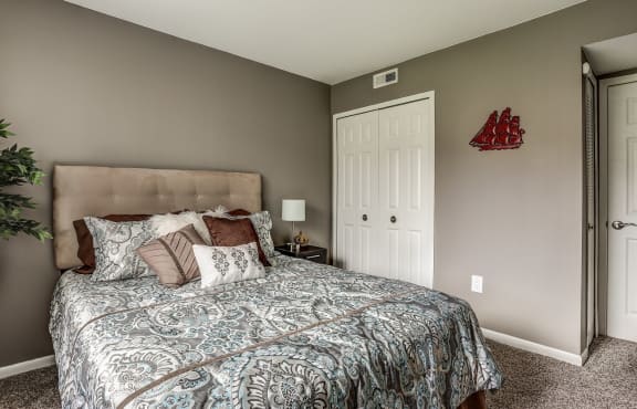 bedroom with tan walls, tan carpeting, and tan headboard. The bed is made in a blue patterned bedding with throw pillows. There are white closet doors to the right of the bed.  