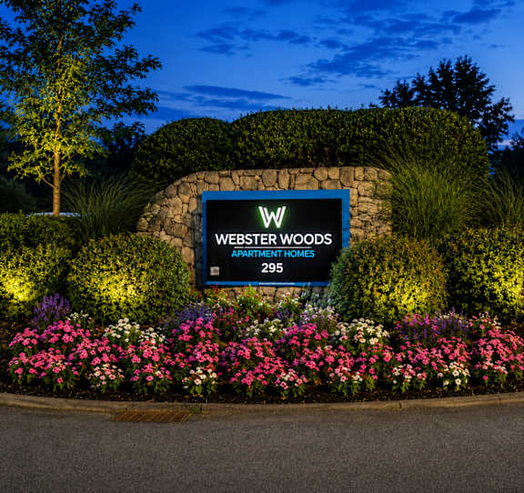 A sign for Webster Woods Apartment Homes is surrounded by flowers and greenery.