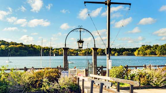 A bench is situated under a metal structure with a sign that reads "PLACE" in front of a body of water.
