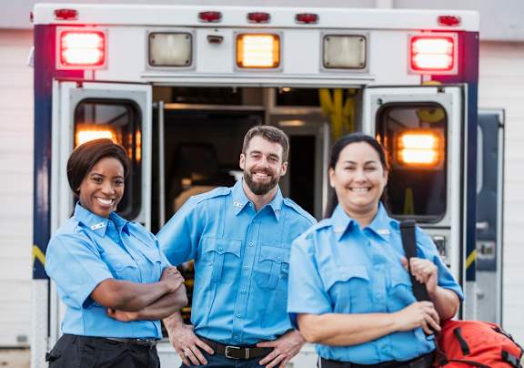 Three people in blue uniforms stand in front of an ambulance.
