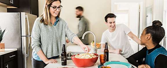 A woman in a grey sweater is serving popcorn to a man and a woman in a kitchen.