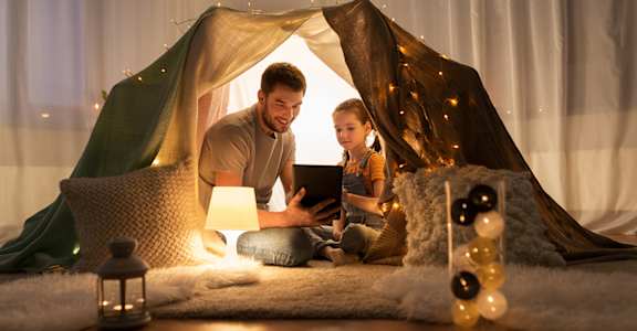 A man and a child are sitting inside a tent-like structure, with the man holding a tablet and the child looking at it.