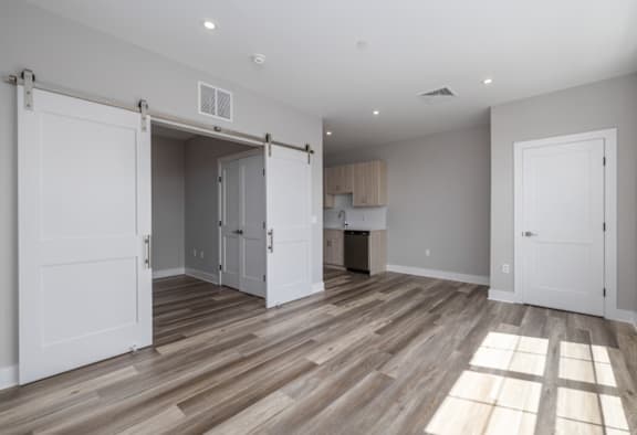 A room with a wooden floor and white walls, with a kitchen area visible through a sliding door.