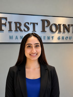 A woman in a black blazer and blue top stands in front of a sign that says "First Pointe Management Group".