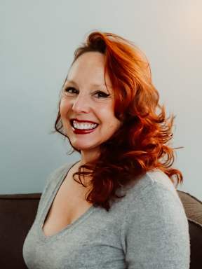 A woman with red hair is smiling and wearing a grey top.