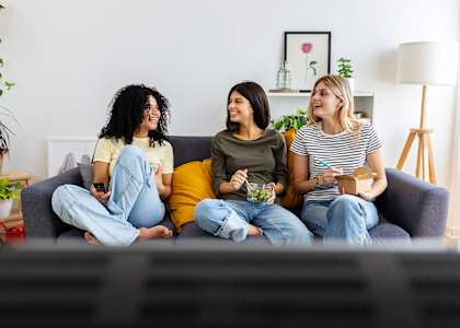 Three women are sitting on a couch, each holding a bowl of salad.