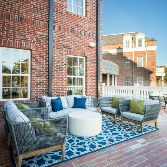 Charming outdoor patio at Tindall Park Apartments featuring cozy wicker seating, colorful cushions, and a stylish blue rug against a classic brick backdrop.