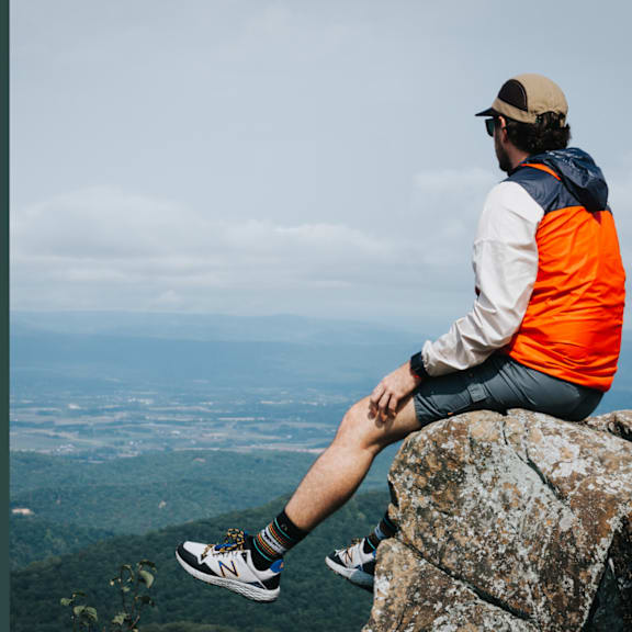 A man in an orange vest and white jacket sits on a rock, overlooking a vast landscape.