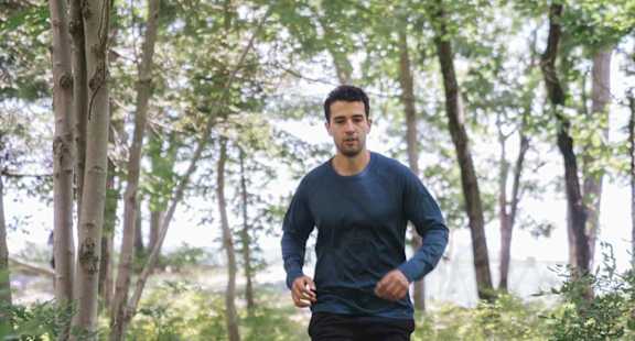 A man in a blue shirt and black shorts is running on a log in a forest.