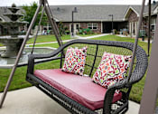 Thumbnail 3 of 11 - A black wicker swing with red cushion and pillows is in the foreground with a house and fountain in the background.