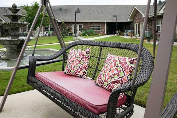 A black wicker swing with red cushion and pillows is in the foreground with a house and fountain in the background.