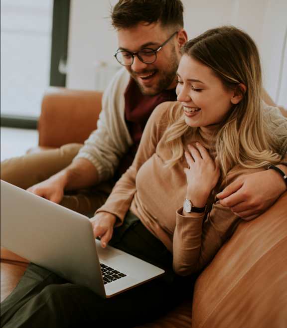 A man and a woman are sitting on a couch, looking at a laptop screen.