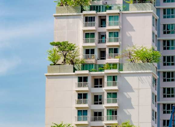 A tall white building with balconies and trees on top.