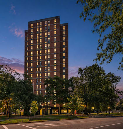 A tall building with many windows is surrounded by trees and has lights on inside.
