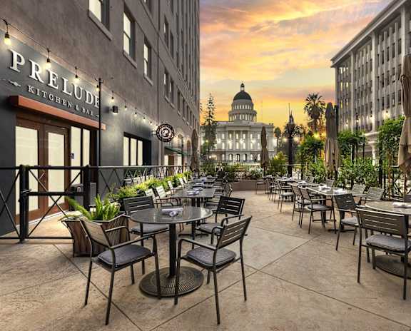 A patio with tables and chairs is set up outside a building with a sign that reads "PRELUDE KITCHEN BAR".