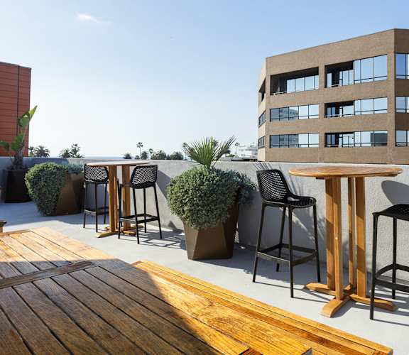 A wooden table is in the foreground of a patio with black chairs and a building in the background.
