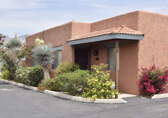 A house with a red tile roof and a brown stucco exterior is surrounded by a variety of plants and flowers at San Xavier Casitas, Arizona, 85716