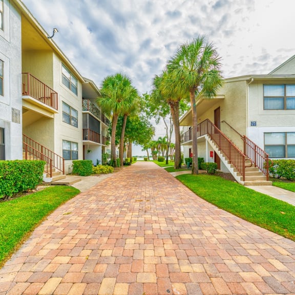 a brick pathway between two apartment buildings with palm trees at Bella Lago Vista, West Palm Beach, FL
