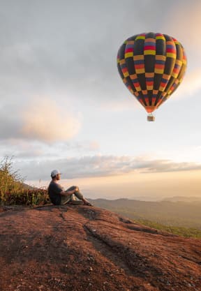 a man sitting on a hill looking at a hot air balloon