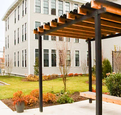 A bench under a pergola with a white building behind it