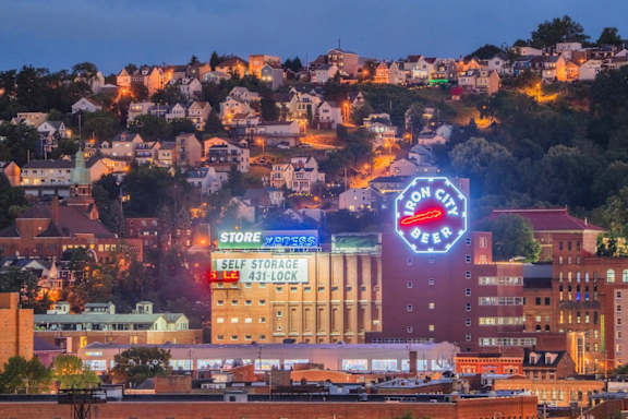 A cityscape at dusk with a prominent building that has a neon sign for "Bron City Beer" and "Store Self Storage 431-Lock.".