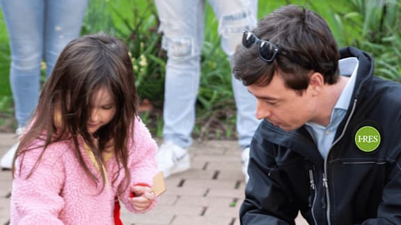 A man in a black jacket with a green patch on the left side of his chest is interacting with a young girl in a pink jacket.