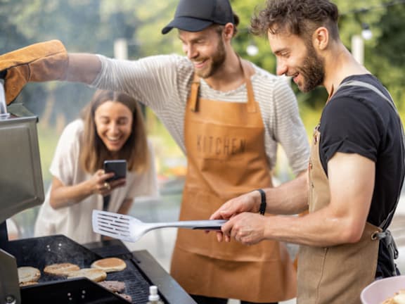 Two men grilling food outdoors with a woman taking a photo.