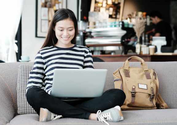 A woman in a striped shirt is sitting on a couch using a laptop.