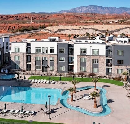 A large swimming pool is surrounded by lounge chairs in front of apartment buildings.