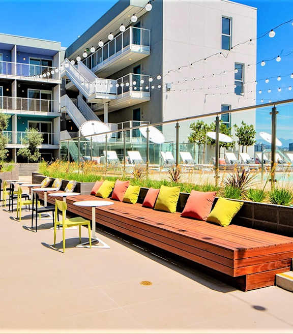 A patio with a long bench and chairs is surrounded by buildings.