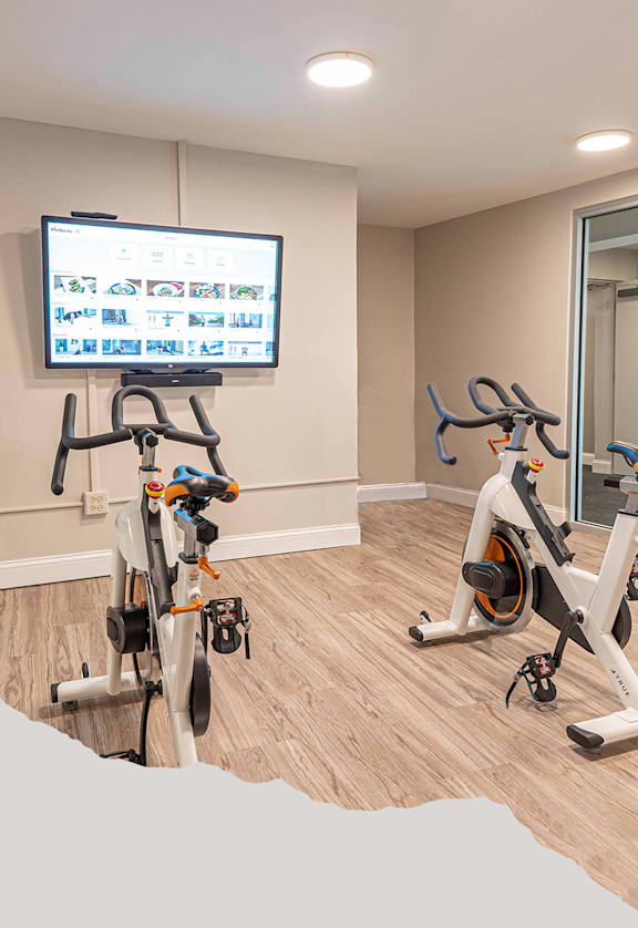 Two exercise bikes are in a room with a TV mounted on the wall.