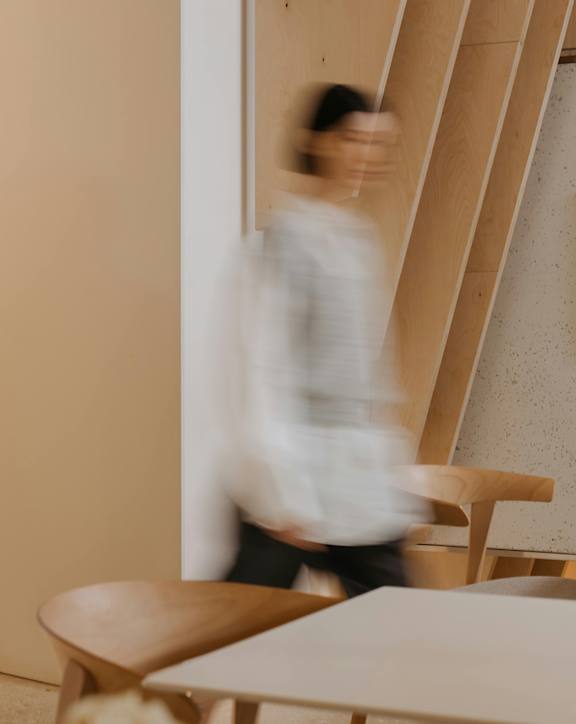 A person in a white shirt is walking past a wooden table and chair.