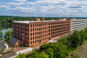 A large red brick building sits next to a body of water. at Montgomery Mill Apartments in Windsor Locks, CT