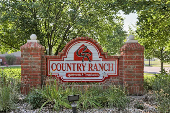 A sign for Country Ranch Apartments & Townhomes is displayed in front of some trees.