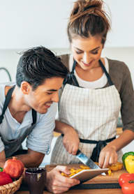 A man and a woman are cooking together, with the woman holding a knife and the man holding a tablet.