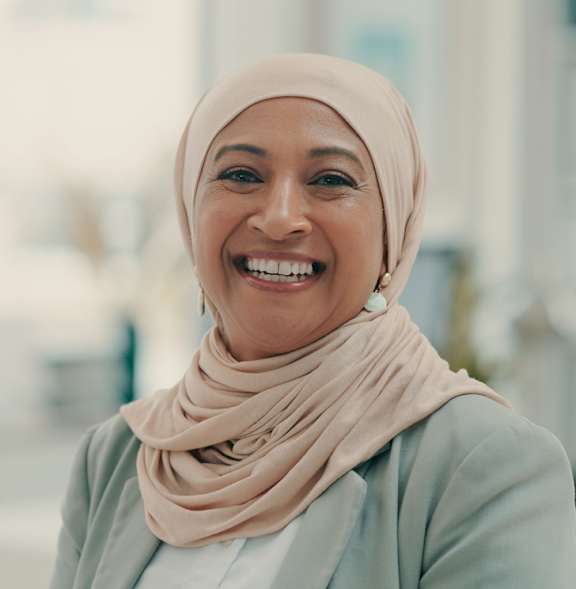 A woman wearing a light-colored hijab smiles at the camera.