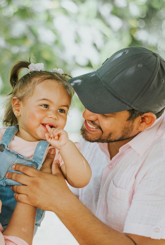 A man in a pink shirt and a black cap is holding a little girl with a white bow in her hair.