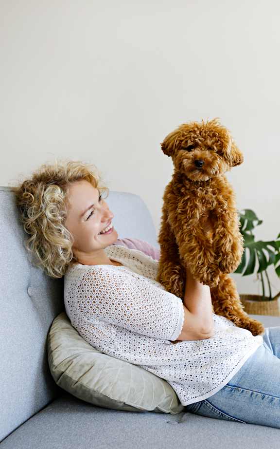 A woman with curly blonde hair is sitting on a couch with a small brown dog on her lap.