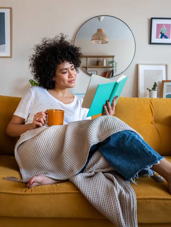 A woman is sitting on a yellow couch, reading a book and holding a mug.
