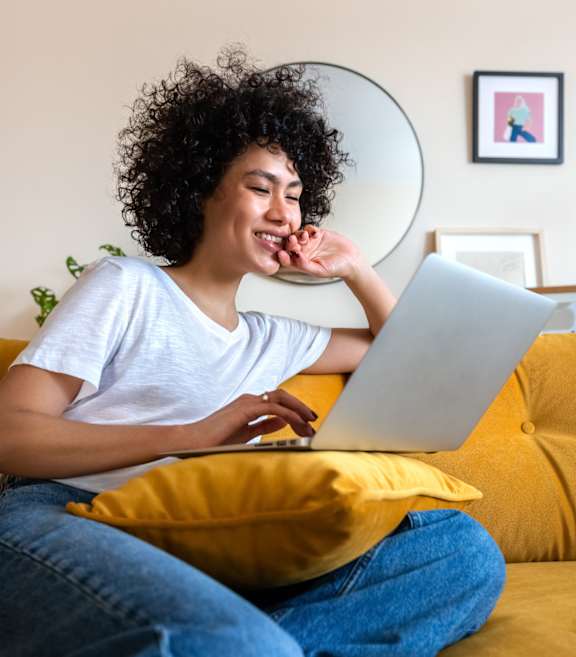 A person with curly hair is sitting on a yellow couch using a laptop.