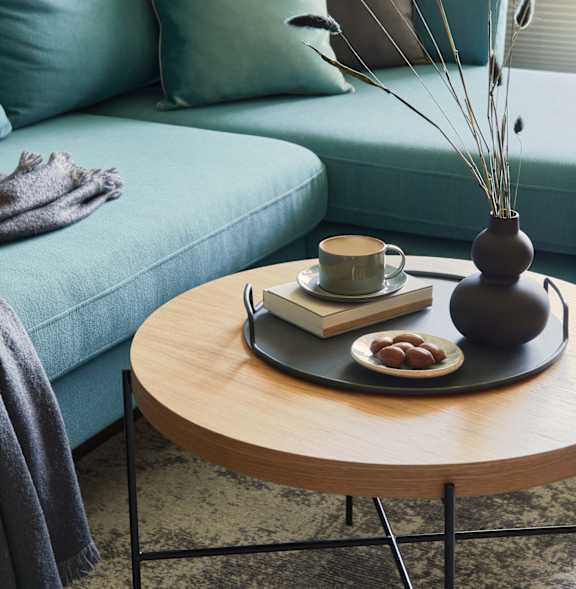 A living room with a blue couch, a wooden table with a vase, a book, a cup, and some fruit.