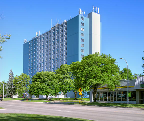 A tall blue building with a grey section at the top is surrounded by trees and a street with cars.