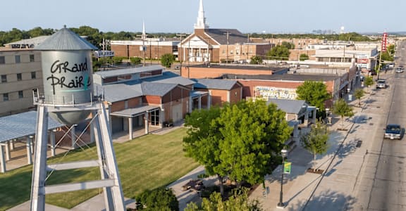 A water tower with Grand Prairie written on it is in the foreground of a city street.