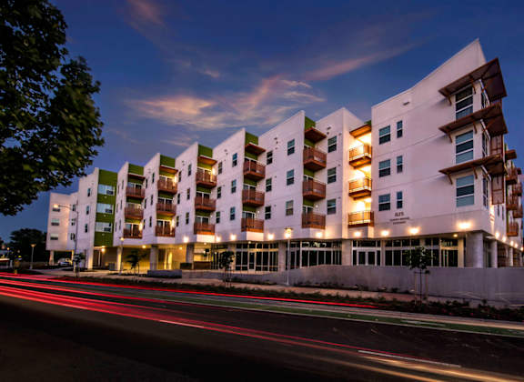 A modern apartment building with a white facade and balconies is illuminated at dusk.